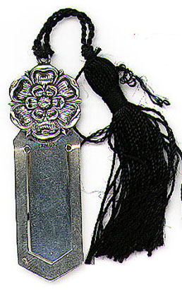 This bookmark was made in Norway. It is marked 830S and has a symbol of a horseshoe surrounding the letters AE. The top of the bookmark is a flower with a black tassel. The date is 1920 - 1930.