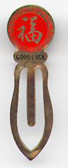 This bookmark was made in China. It is made of tin and enamel paint and is red with a Chinese symbol on the top. Written underneath the symbol is "Good Luck". The date is 1970 - 1980.  