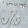 This bookmark was made in the US by an unknown manufacturer. It is marked only sterling. It depicts two cherub heads, a girl and a boy where the boy is kissing the girl. The heads are above two wings. The top blade is a cut out heart shape. The date is 1900 - 1910.