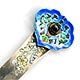 This bookmark was made in China. It is hallmarked with a Chinese symbol. The top has blue enamel with green and red flowers surrounding a brownish stone. The blade has inscribed enameled flowers.