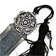 This bookmark was made in Norway. It is marked 830S and has a symbol of a horseshoe surrounding the letters AE. The top of the bookmark is a flower with a black tassel. The date is 1920 - 1930.